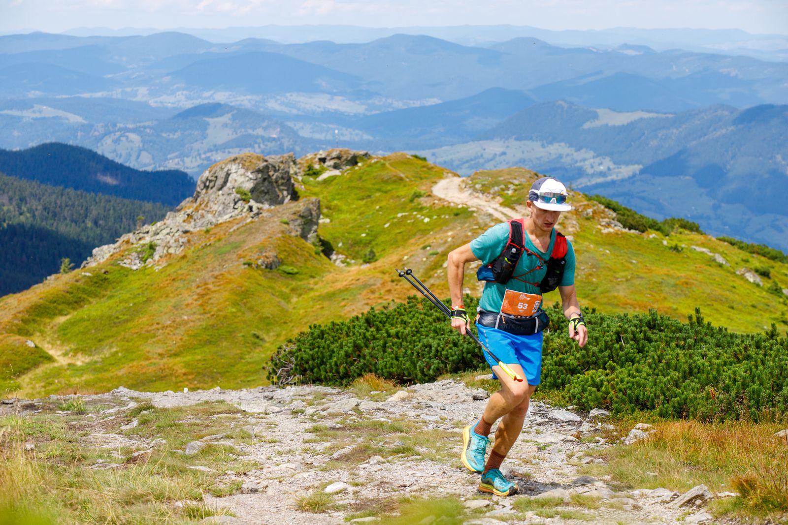 Un coureur de trail porte un gilet d’hydratation et des bâtons pliés sur un sentier de montagne rocheux.